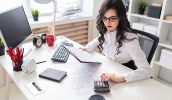 A young girl is sitting at the office desk and is blessing on the calculator.