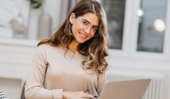 Photo of lovely white woman in glasses using computer with charming smile. Indoor portrait of stunning student with curly hair studying with laptop..