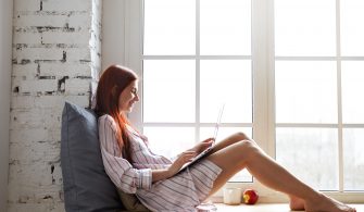 Sideways shot of cheerful teenage girl in striped shirt relaxing by window, speaking to friend via online video chat, using laptop. Pretty woman sitting barefooted on windowsill with portable computer