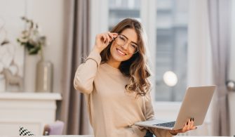 Cute girl in beige shirt touching glasses and holding laptop with smile. Indoor photo of beautiful female student preparing for lesson with computer at home..