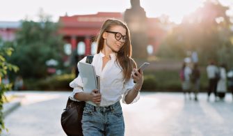 Smiling brunette student wearing glasses and shirt walking outdoor  with laptop and backpack and looking in smartphone in sunlight on background of university