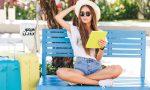 Cute girl sitting on a blue bench and playing on a tablet in yellow case. She wears denim shorts, white t-shirt, dark sunglasses, white sneakers and straw hat. She sits with two suitcases next to her