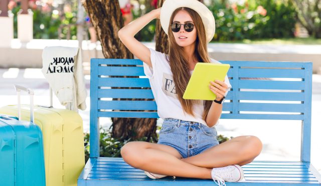 Cute girl sitting on a blue bench and playing on a tablet in yellow case. She wears denim shorts, white t-shirt, dark sunglasses, white sneakers and straw hat. She sits with two suitcases next to her