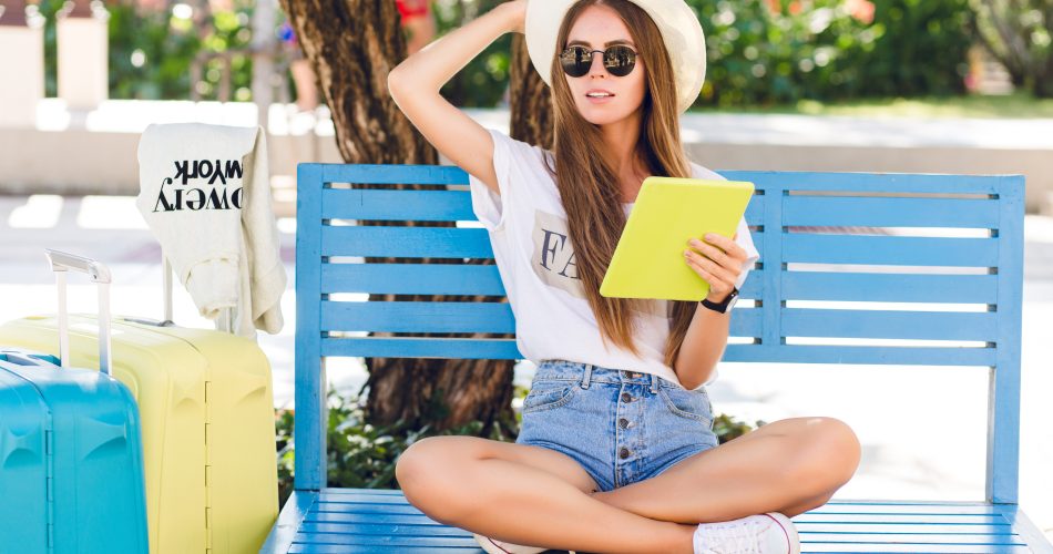 Cute girl sitting on a blue bench and playing on a tablet in yellow case. She wears denim shorts, white t-shirt, dark sunglasses, white sneakers and straw hat. She sits with two suitcases next to her