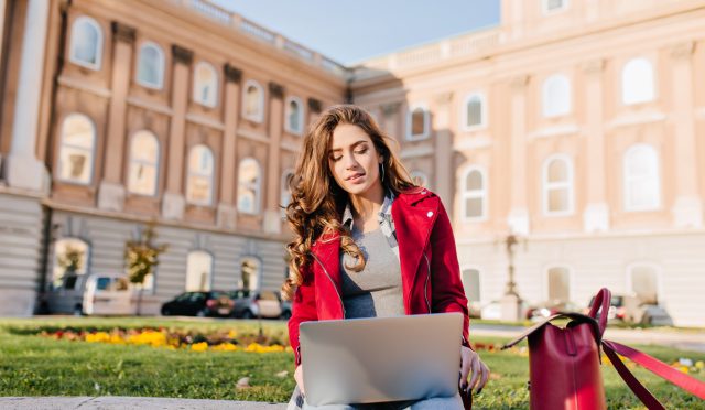 Outdoor portrait of serious curly female student sitting with laptop on the ground. Busy brunette girl in red jacket working with computer in front of college in warm day..