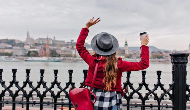 Portrait from back of stylish young woman in checkered shirt waving hands on city background. Excited long-haired female model enjoyng river views in cold day..